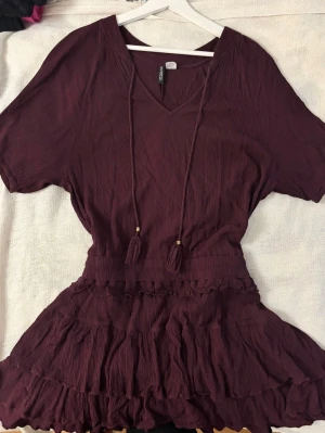 Vinröd klänning med guld detaljer . H&M strl M - Elegant vinröd klänning från H&M i superbekvämt material. V-ringad med trekvartsärm, fina tassel-detaljer och dekorativa guldsnören. Volangkjol som ger en fin och feminin passform. Använd fåtal gånger och i väldigt fint skick. ❤️ 