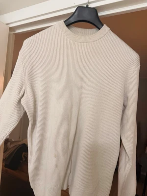 Beige ribbstickad tröja - Säljer en stilren beige ribbstickad tröja med rund hals och långa ärmar. Tröjan har en relaxed passform och breda muddar vid ärmslut och nederkant. Perfekt att matcha med jeans eller kjol för en enkel och clean look.3 fläckar nertill som kan gå bort i tvätt, därav priset