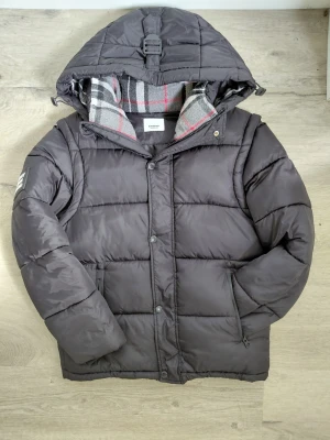 Svart pufferjacka från Burberry - Säljer en svart pufferjacka från Burberry med avtagbar huva och klassiskt rutigt foder. Jackan har en patch med 'BURBERRY LONDON ENGLAND' på ärmen, dragkedja och knappar framtill samt två sidofickor. Perfekt för kalla vinterdagar.
