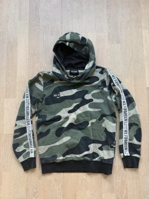 Camouflage hoodie med tryck - Säljer en grön och svart camouflage hoodie med huva och svarta muddar. På ärmarna finns vita band med text.