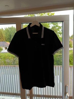 Svart pikétröja från BOSS - Klassisk svart pikétröja från BOSS med vit logga på bröstet. Tröjan har krage med vita ränder och matchande detaljer på ärmsluten. Perfekt till jeans eller shorts för en clean och stilren look. Hör av er vid fler frågor!