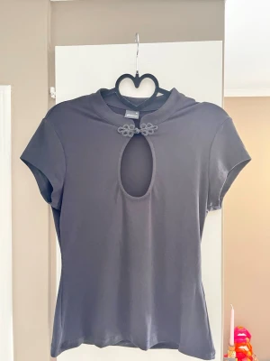 Svart topp med cut-out och dekor - Svart topp med korta ärmar och unik cut-out framtill vid halsen, dekorerad med två små blommiga detaljer. Mjuk och stretchig känsla, troligen i bomull eller polyester. Snygg och enkel att styla till olika looks.