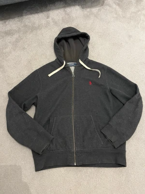 Mörkgrå hoodie med dragkedja - Snygg mörkgrå hoodie från Polo Ralph Lauren med dragkedja framtill och klassisk röd logga broderad på bröstet. Hoodien har vita snören i huvan, två fickor fram och ribbade muddar. Perfekt för en avslappnad och stilren look.