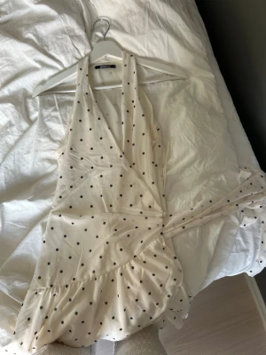 Prickig omlottklänning från Gina Tricot - Söt kort omlottklänning från Gina Tricot i ljus beige med svarta prickar. Klänningen har volangkant nertill, knyt i midjan och djup v-ringning. Perfekt för sommarens varma dagar och har en luftig, ärmlös design. Den är inte så v ringad som den ser ut på bilden utan du kan välja djupet på v ringning själv när du väl bär den. Använd 1 gång 