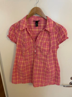 Rosa-rutig blus med korta ärmar, mamma blus för gravida, bomull, stl L - Söt blus i rosa och orange-rutigt mönster med korta puffärmar och krage. Blusen har knappar framtill och två bröstfickor. Tillverkad i ett lätt och luftigt tyg, perfekt för varma dagar. Passar dig som gillar färg och en avslappnad stil. mamma blus för gravida, bomull, stl L