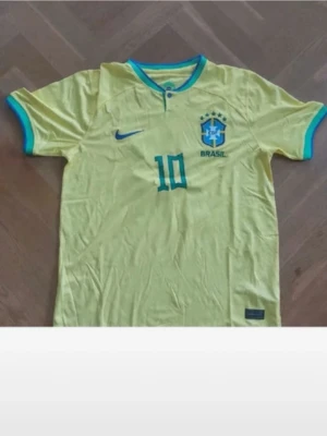 Gul Brasilien t-shirt med nummer 10 - Snygg gul Brasilien t-shirt med blå detaljer och nummer 10 på bröstet. T-shirten har korta ärmar, rund halsringning med blå kant och tryckt Brasilien-emblem. Perfekt för dig som älskar fotboll och vill visa ditt stöd för landslaget.