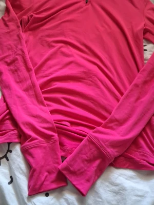 Rosa långärmad träningströja - Säljer en starkt rosa långärmad träningströja med tumhål vid ärmsluten och halv dragkedja vid halsen. Tillverkad i mjukt och stretchigt material som passar perfekt för löpning eller gym. Snygg och funktionell för aktiva dagar.