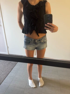 Svart volang topp med struktur - Säljer en svart ärmlös topp med volangdetaljer framtill och krage. Tyget har en snygg struktur och toppen är croppad. Perfekt för dig som vill ha en trendig och unik look. Materialet känns som polyester.