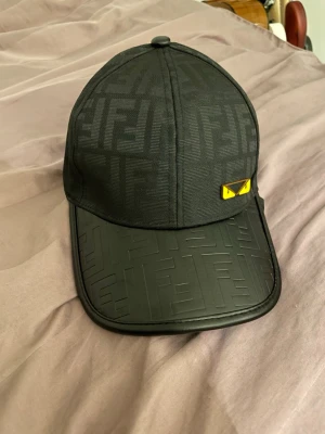 Svart Fendi keps - Snygg svart keps från Fendi med det klassiska FF-mönstret över hela kepsen och skärmen. Skärmen är i ett glansigt material med präglat mönster och på sidan finns en gul detalj med ögonmotiv. Perfekt för dig som gillar exklusiva accessoarer.
