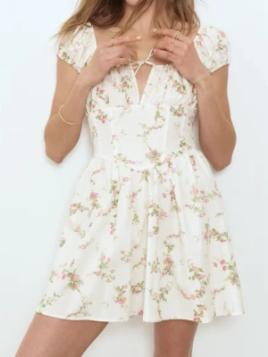 Vit blommig kort klänning med puffärm - Söt kort klänning i vit bas med små rosa och gröna blommor. Klänningen har puffärmar, markerad midja och en liten knytning vid urringningen. Tillverkad i ett lätt, mjukt tyg som ger en romantisk känsla.