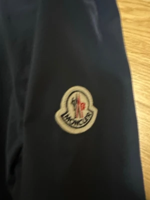Mörkblå vindjacka från Moncler - Snygg mörkblå vindjacka från Moncler med klassisk logotyp på ärmen. Jackan har en minimalistisk design och är tillverkad i ett lätt och tåligt material som passar perfekt för blåsiga dagar. Stängs med tryckknappar och har en stilren look. Storlek 3=L