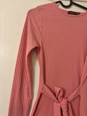 Rosa ribbad klänning med knyt - Säljer en rosa ribbad klänning från Boohoo med lång ärm och knyt i midjan. Klänningen har en kroppsnära passform och är tillverkad i ett mjukt, stretchigt material. Perfekt för dig som gillar enkel men snygg stil.