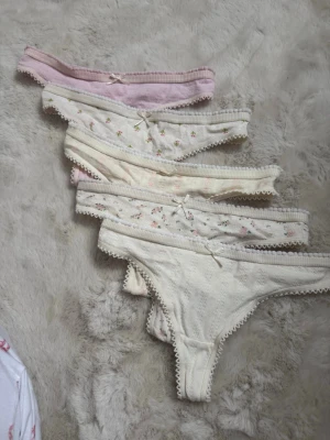 Söta spetskantade trosor i pastell - Fem par trosor i mjuk bomull med spetskant och små rosetter framtill. Färgerna är ljusrosa och olika nyanser av vitt, några med blommigt mönster i rosa och grönt. Klassisk trosa-modell med romantisk känsla och fina detaljer.