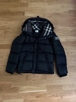 Svart Burberry jacka - Snygg svart dunväst från Burberry med avtagbara ärmar och huva. Västen har klassiskt rutigt foder i huvan, dragkedjor på fickorna och tryckknappar framtill. Perfekt för lager-på-lager och streetstyle.