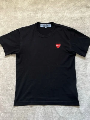 Cdg Svart t-shirt med rött hjärta - Snygg svart t-shirt från Comme des Garçons Play med det ikoniska röda hjärtat med ögon broderat på bröstet. Klassisk passform och rund halsringning. Perfekt till jeans eller kjol för en clean och trendig look. Storlek S 