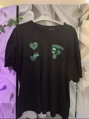 Svart t-shirt med gröna tryck - Cool svart t-shirt med gröna tryck framtill, bland annat ett hjärta, alien och texten 'Happiness'. T-shirten har rund hals och korta ärmar. Perfekt för dig som gillar grafiska detaljer och en avslappnad stil.