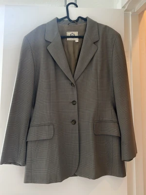 Militärgrön rutig kavaj med tre knappar - Stilren grå kavaj med diskret rutmönster och tre knappar framtill. Klassisk krage och två fickor med lock. Perfekt för en dressad look och enkel att matcha med olika outfits. Fodrad insida för extra komfort.