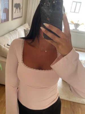 Ljusrosa topp med knyt och spets - Supersöt ljusrosa topp med långa ärmar, fyrkantig ringning och spetsdetaljer vid halsen. Toppen har öppen rygg med knytband som ger en extra snygg touch. Perfekt för dig som gillar feminina detaljer och trendig stil.