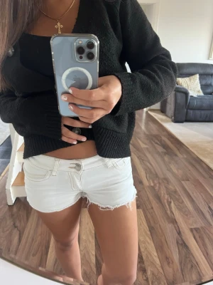 Vita jeansshorts med detalj på fickorna  - Säljer ett par vita jeansshorts med detaljer på fickorna och slitna detaljer. Shortsen har klassisk femficksdesign, bälteshällor och snygga broderade detaljer på bakfickorna. Perfekta för varma dagar och enkel att matcha med allt.