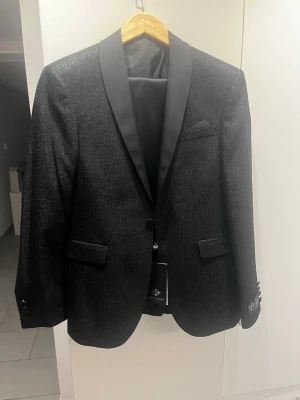 Svart glittrig kavaj och byxa från Marco Menti - Snygg svart kavaj från Marco Menti med glittrig finish och slät satinrevers. Kavajen har två fickor med lock, bröstficka och dekorativa knappar vid ärmslutet. Även en byxa. Perfekt för dig som vill sticka ut med stil. Aldrig använd!! Pris kan diskuteras. 