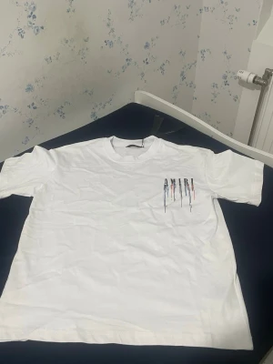 Vit Amiri t-shirt med färgstänk - Säljer en vit t-shirt från Amiri med tryck i svart, blått och rött både fram och bak. T-shirten har korta ärmar och en relaxed passform. Materialet är mjuk bomull och trycket ger en cool streetwear vibe.Vi kan komma överens med priset