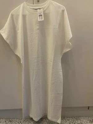 Vit t-shirtklänning från Kappahl - Enkel och stilren vit t-shirtklänning från Kappahl i mjuk bomull. Klänningen har rund halsringning, korta ärmar och rak passform. Perfekt för en avslappnad och trendig look.
