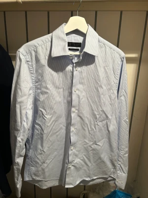 Ljusblå smalrandig skjorta - Klassisk ljusblå skjorta med tunna vita ränder och knappar framtill. Skjortan har lång ärm, normal passform och en stilren krage. Perfekt för en clean och fräsch look.         PRIS KAN DISKUTERAS 