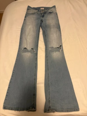 Ljusblå utsvängda jeans med hål - Säljer ett par ljusblå jeans med utsvängda ben och slitna hål på båda knäna. Jeansen har klassisk femficksdesign och är tillverkade i denim med en snygg tvättad look. Perfekta för en trendig och avslappnad stil.