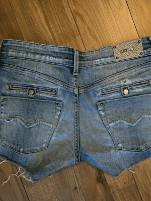 Blå jeansshorts från Replay - Snygga ljusblå jeansshorts från Replay med råa kanter och slitna detaljer. Shortsen har bakfickor med knapp och dekorativa sömmar samt bälteshällor. Hittar ingen storlek men skulle säga S/XS. Midjemåttet 78 cm