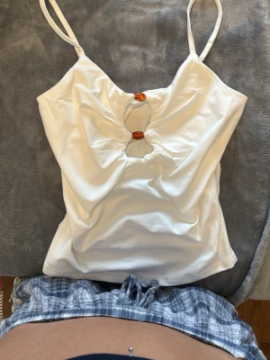 Vit linnetopp med cut-out och pärldetaljer - Snygg vit linnetopp med smala axelband och två bruna pärldetaljer framtill som skapar en cut-out effekt.  Märket är oklart