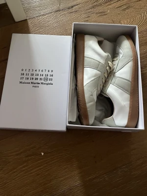 Maison Margiela vita sneakers - Maison Margiela sneakers i vitt läder med grå mockadetaljer och klassisk gummisula. Snygg minimalistisk design med låga skaft och vita snören. Perfekta för dig som gillar stilrena och trendiga sneakers.