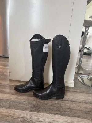 Svarta höga ridstövlar i läder - Snygga svarta ridstövlar i läder med klassisk design och högt skaft. Stövlarna har rundad tå, låg klack och dekorativa knappar upptill. Perfekta för dig som gillar stilrena och tidlösa boots till ridning.           Aldrig använda 