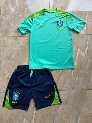 Brasilien fotbollströja och shorts Nike - Säljer ett Brasilien fotbollskit från Nike med ljusgrön t-shirt och mörkblå shorts. Tröjan har gröna axelpartier, gul Nike-logga och Brasilien-emblem på bröstet. Shortsen är mörkblå med gröna detaljer, gul Nike-logga och Brasilien-märke. Perfekt för träning eller match!