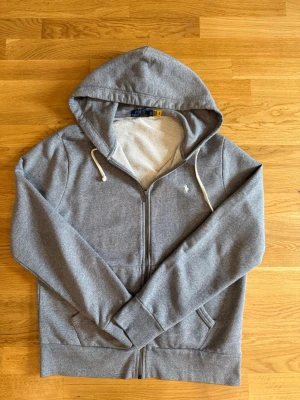 Grå hoodie från Polo Ralph Lauren - Snygg grå hoodie med dragkedja från Polo Ralph Lauren. Klassisk design med vit broderad logga på bröstet, huva med vita snören och två fickor framtill. Tillverkad i mjukt bomullsmaterial som känns skönt mot huden.