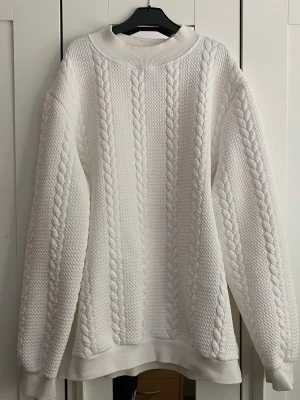Vit kabelstickad tröja - Säljer en oversized vit stickad tröja med klassiskt kabelstickat mönster. Tröjan har rund halsringning, långa ärmar och ribbade muddar vid ärmslut och nederkant. Perfekt för lager-på-lager och en clean look.