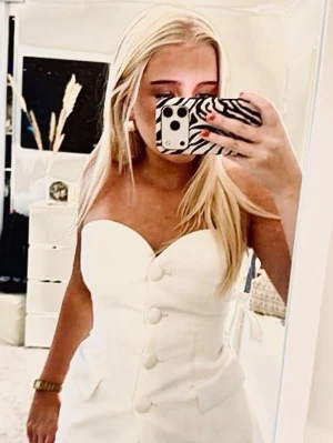 Vit bandeau topp med stora knappar - Säljer en vit bandeau topp med stora dekorativa knappar framtill och två fickdetaljer. Toppen är axelbandslös och har en figurnära passform som ger en clean och trendig look. Perfekt att styla med jeans eller kjol för en snygg outfit. Helt oanvänd 