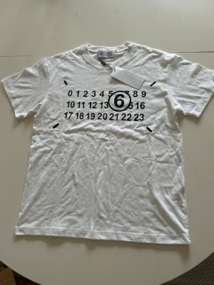 Vit t-shirt med tryck från Margiela - Säljer en vit t-shirt från Maison Margiela med det ikoniska siffertrycket och en stor svart 6:a på bröstet. T-shirten har rund hals och korta ärmar. Perfekt för dig som gillar minimalistisk och stilren design.
