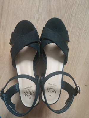 Svarta sandaler med klack från VOX stl37 - Stilrena svarta sandaler från VOX med breda korslagda remmar och justerbart spänne i silver. Klackarna är blockformade och materialet ser ut att vara veganskt mockaimitation. Perfekta för dig som vill ha en trendig och bekväm look. Använda en gång.