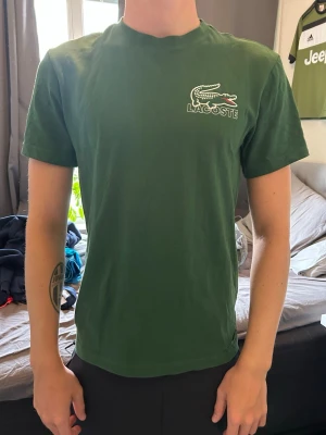 Grön Lacoste t-shirt med tryck - Snygg grön t-shirt från Lacoste med klassisk krokodil-logga och text på bröstet samt stort tryck med Lacoste och krokodil på ryggen. T-shirten har rund hals och korta ärmar, perfekt för en avslappnad stil.