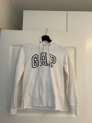 Vit zip hoodie från GAP - vit hoodie från GAP med dragkedja framtill och stor logga i svart på bröstet. Tröjan har huva med dragsko och två fickor framtill. Lite nopprig se bild