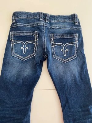 Blå bootcut jeans med slitningar - Säljer ett par blå bootcut jeans med snygga broderade bakfickor. Jeansen har vit kontrastsöm längs sömmarna och en klassisk femficksmodell. Perfekta för dig som gillar en avslappnad och trendig stil.Dom är från Ginatricot Young och är i storlek 158