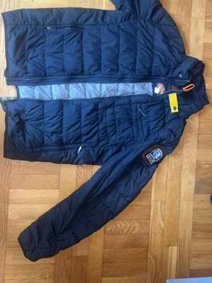 Mörkblå dunjacka med patch - Snygg mörkblå dunjacka med quiltad design och dragkedja framtill. Jackan har en tydlig patch på ärmen med texten 'THAT OTHERS MAY LIVE' och 'P.J.S ANCHORAGE'. Fodrad insida och praktiska fickor med dragkedja. Perfekt för kalla vinterdagar.