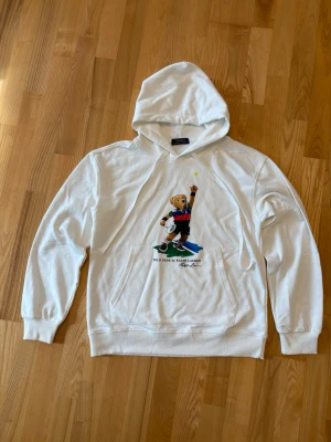 Vit hoodie Polo Bear Ralph Lauren - Vit hoodie från Ralph Lauren med Polo Bear-tryck framtill där björnen spelar tennis. Klassisk huva med dragsko och stor magficka. Hoodien har ribbade muddar vid ärmslut och nederkant. Perfekt för dig som gillar sportig och lekfull stil. Äkta och nästan inte använd. Nypris 1500.