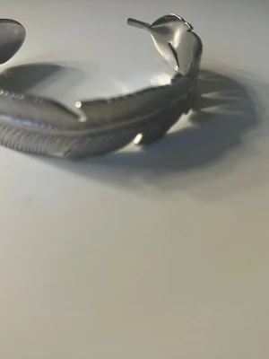 Silvrigt armband med fjäderdesign - Snyggt armband i silverfärgad metall format som en fjäder. Detaljerad gravyr som ger en naturinspirerad look. Perfekt för dig som gillar unika accessoarer med boho-vibe. Justerbar storlek tack vare öppningen.