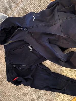 Race marine tracksuit/mjukiskläder - Mysigt svart loungewear-set med zip-hoodie och matchande byxor. Hoodien har dragsko och tryck på bröstet, byxorna har resår i midjan och broderad text. Perfekt för chill dagar hemma.