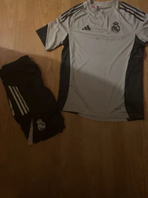 Real Madrid träningsset Adidas - Säljer ett snyggt Real Madrid träningsset från Adidas. Setet består av en ljusgrå t-shirt med mörkgrå detaljer och matchande shorts. Båda plaggen har klubbens logga och klassiska Adidas-ränder. Perfekt för fotbollsträning eller gymmet.