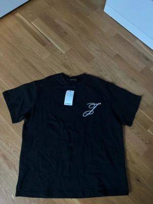 Jacquemus t-shirt  - Säljer en svart t-shirt från Jacquemus med vit logga på bröstet och stort Jacquemus-tryck på ryggen. Klassisk rund hals och korta ärmar. Tillverkad i mjuk bomull för skön känsla och enkel stil.
