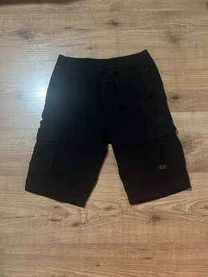 Svarta cargoshorts med fickor - Svarta cargoshorts med flera praktiska fickor och snörning i midjan. Perfekta för en avslappnad stil och har en loose passform. Shortsens material känns mjukt och bekvämt, troligen bomull eller polyester. stoleken ser ni på bilden men det är stolek 146 