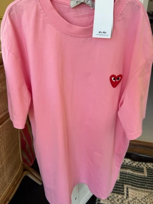 Rosa Comme des Garçons PLAY t-shirt - Säljer en rosa t-shirt från Comme des Garçons PLAY med det ikoniska röda hjärtat broderat på bröstet. T-shirten har rund hals, korta ärmar och är gjord i mjuk bomull. Perfekt för dig som gillar streetwear och vill sticka ut lite extra.