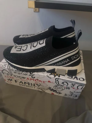 Dolce & Gabbana svarta sneakers - Svarta slip-on sneakers från Dolce & Gabbana med glittrande stenar över hela ovandelen och stora vita logotexter på ovansidan. Skorna har en chunky vit och svart sula samt dragflikar bak. Snygg streetstil med exklusiv känsla. Storlek 43 utmärkt skick.
